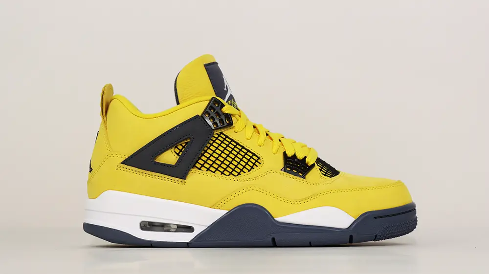 A front view of the outer side of the right shoe of the Jordan 4 Retro ‘Lightning’ (2021), featuring black mesh with breathable perforations and a predominantly yellow suede upper.”