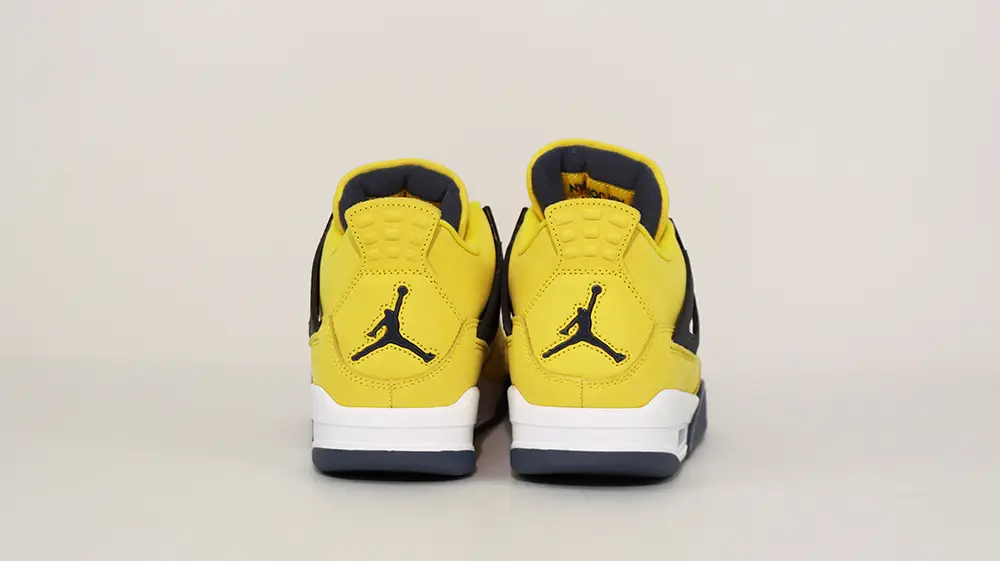 A rear view of both shoes of the Jordan 4 Retro ‘Lightning’ (2021), showcasing the overall yellow leather upper and the metallic black Jordan logo on the heel.