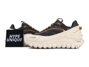 Dark and beige Moncler sneakers with thick, textured soles are displayed on a white surface. The shoes have visible branding, including "GORE-TEX" and "Vibram." A black card with "HYPE UNIQUE" stands beside the sneakers.