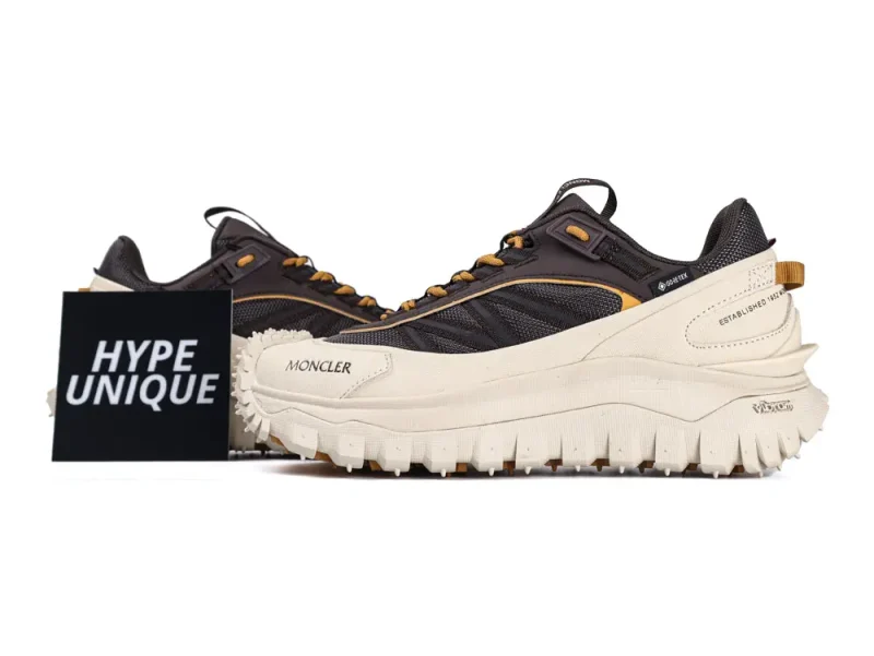 Dark and beige Moncler sneakers with thick, textured soles are displayed on a white surface. The shoes have visible branding, including "GORE-TEX" and "Vibram." A black card with "HYPE UNIQUE" stands beside the sneakers.