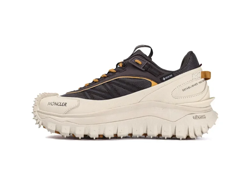 Athletic shoe with a chunky, textured sole and a blend of beige and black colors. Features include Gore-Tex material, Moncler branding, and a thick tread pattern for traction. Orange accents and heel loops provide additional design elements.