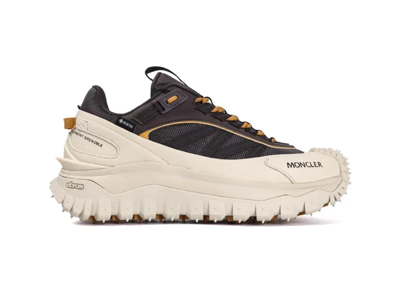 Rugged sneaker with a black mesh upper, beige leather paneling, and a chunky beige sole. Features include Gore-Tex branding, yellow lace highlights, and pronounced text labeled "MONCLER" and "Vibram." Designed for outdoor performance.