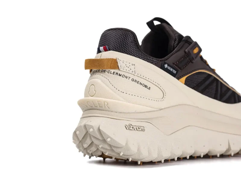 Close-up of a black and beige sneaker featuring Gore-Tex technology, detailed stitching, and textured sole. Labels include Moncler and Vibram. The shoe has a pull tab and design elements for outdoor use.