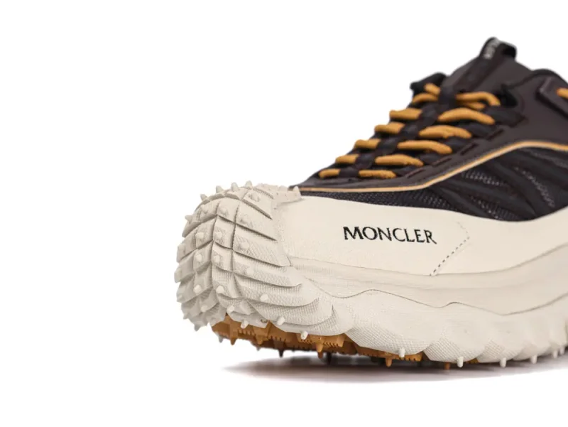 Close-up of a sneaker focusing on the toe and side, featuring a textured white sole with a rugged pattern. The shoe is black with brown laces, and has "Moncler" printed on the side. The design includes intricate detailing on the upper part, combining mesh and fabric materials.