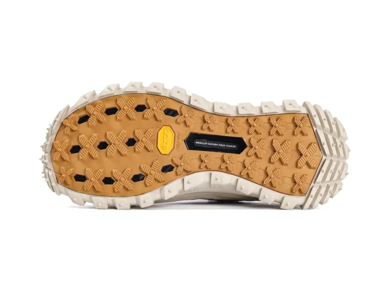 Sneaker sole with rugged traction pattern, featuring hexagonal and diamond-shaped lugs in beige and black colors. The design highlights grip and durability, with a visible brand logo and "Moncler Carbon Fiber Chassis" text on a label.