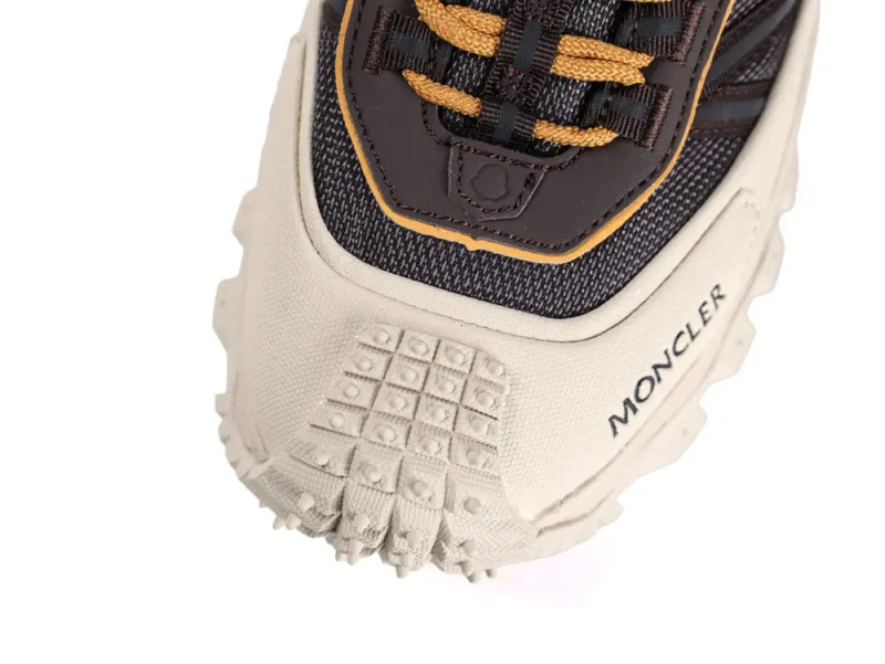 Close-up of a sneaker's front, featuring beige textured rubber sole, dark fabric with yellow and black laces, and "Moncler" branding on the side.