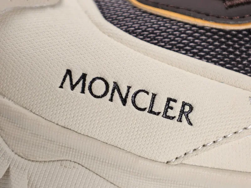 Close-up of a sneaker featuring the brand name "Moncler" on textured white material, with dark fabric and yellow accent stitching in the background.