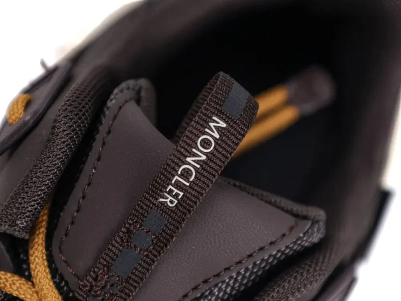 Close-up of a brown Moncler sneaker's opening, showcasing textured black and brown fabric, leather detailing, and a black pull tab with white Moncler text. Brown laces are visible in the background.