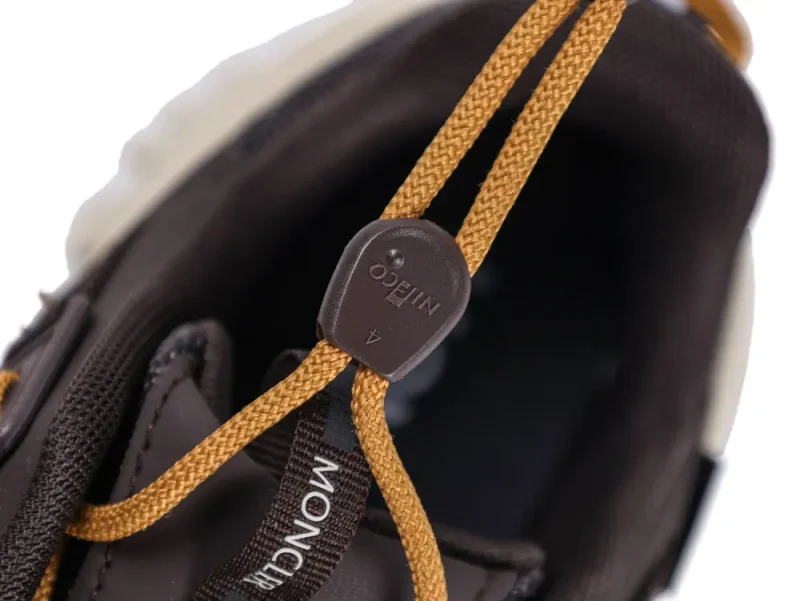 Close-up of a brown shoe with mustard laces featuring a toggle lock. The word "MONCLER" is partially visible on the tongue. The image highlights the lace mechanism and shoe's interior.