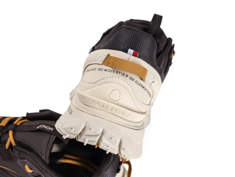 Close-up of a sneaker's heel, featuring a combination of dark brown and tan materials. The heel displays branding details, including text and a logo. The shoe's sole is textured and prominently detailed.