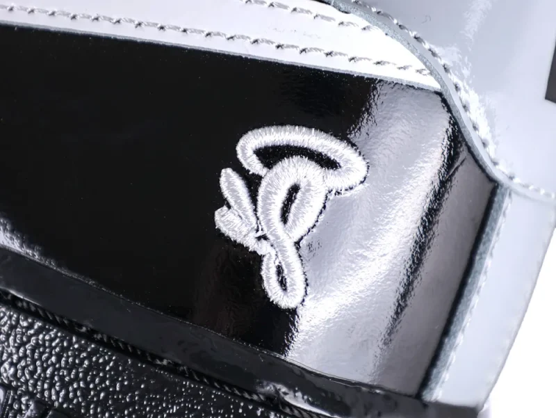Close-up of the white embroidered Syna World "C" logo on the black patent leather heel.